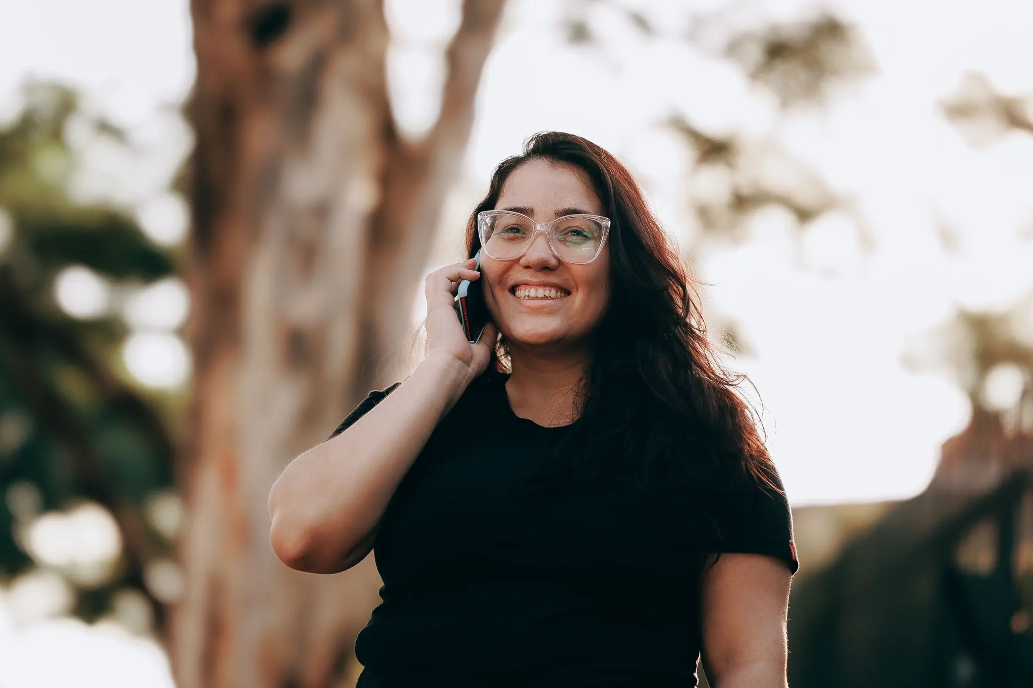 A woman in glasses laughing as she talks on the phone under the trees