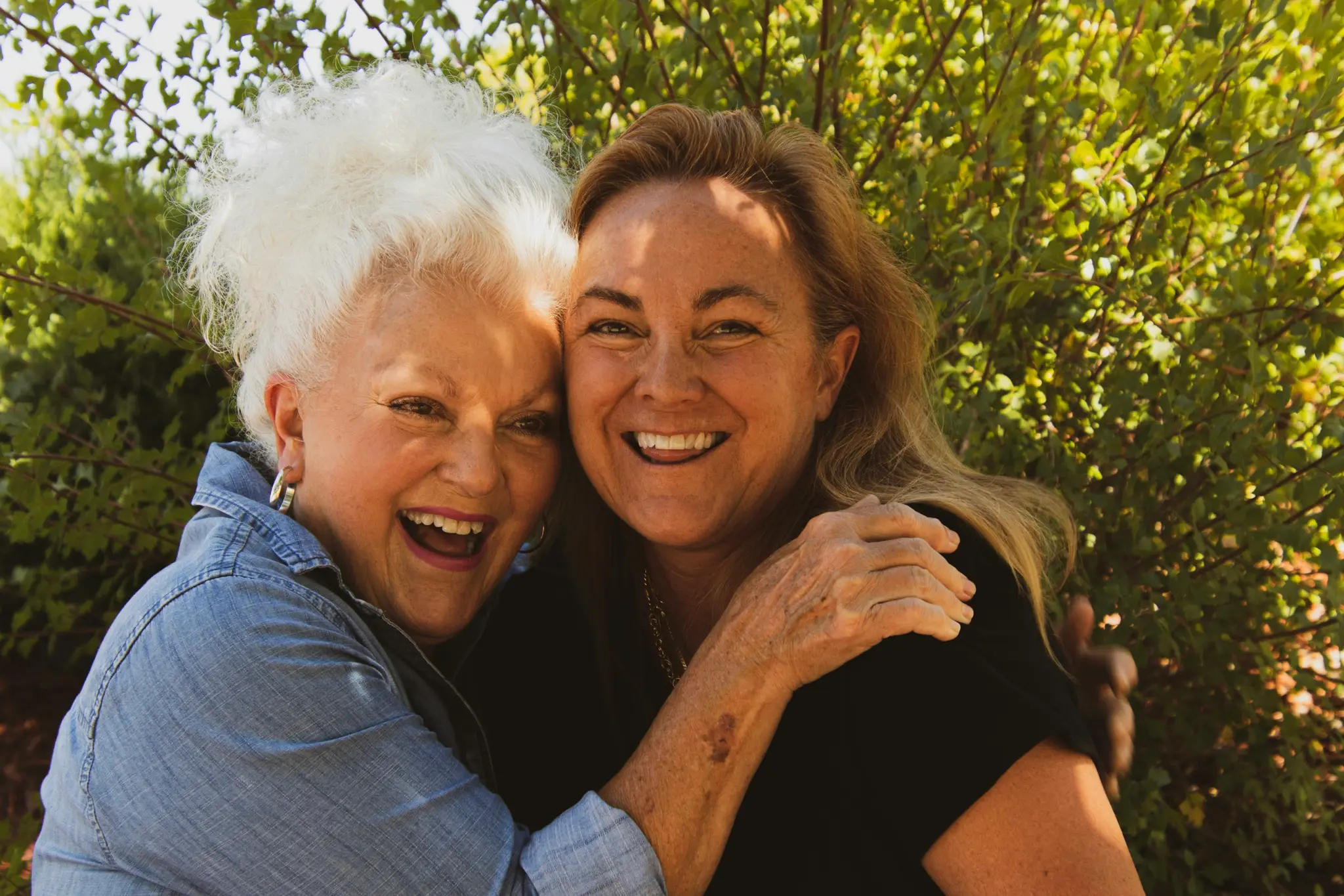 A mother and her grown daughter laughing cheek to cheek outside