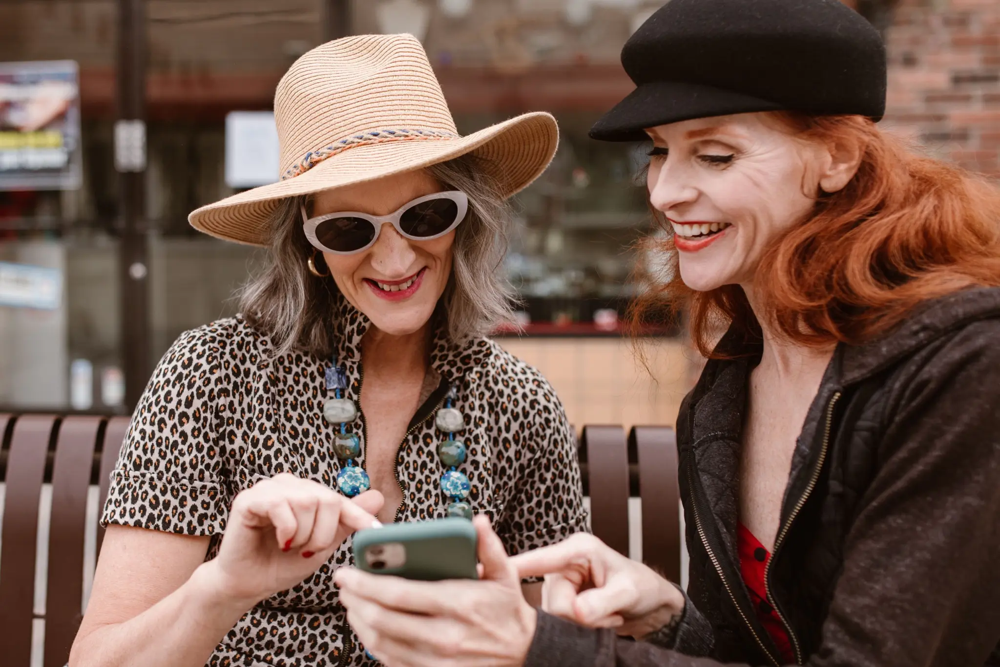 Two women laughing together over a phone on a city bench
